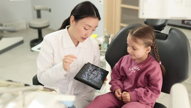 Medicine, pediatric dentistry and oral care concept. Female smiling asian dentist showing tablet pc computer to happy kid patient, caucasian preschool girl at modern dental clinic.