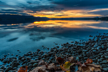 Fototapeta na wymiar Bodenseeufer im Herbst mit Blättern und Steinen im Vordergrund zum Sonnenuntergang 