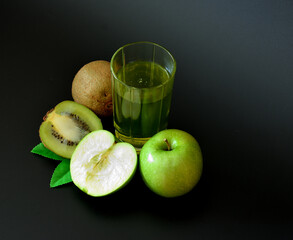 Fruit juice in a tall glass on a black background, next to ripe fruits of kiwi and a green apple.