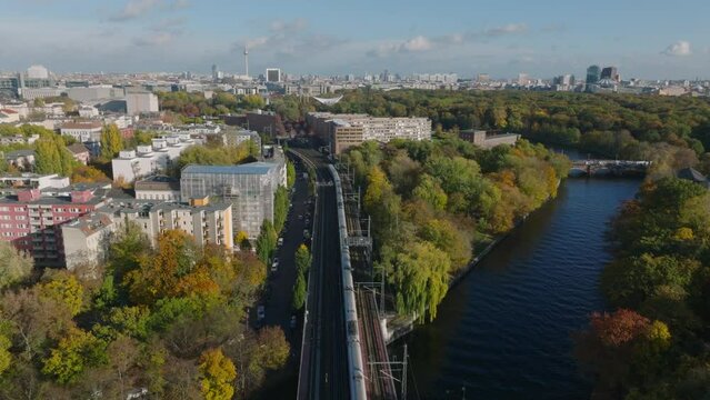 Aerial view of passenger train passing on bridge over river. Rising reveal of large Tiergarten park and cityscape with famous Fernsehturm. Berlin, Germany