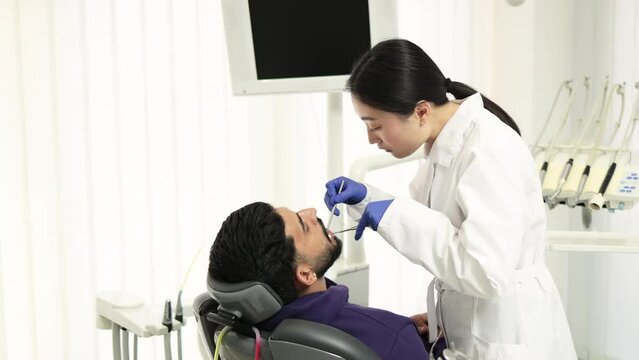 Side view of young pretty asian doctor in white uniform and gloves, making examination or curing caries and toothache for her male patient, attractive bearded man sitting in dental chair.
