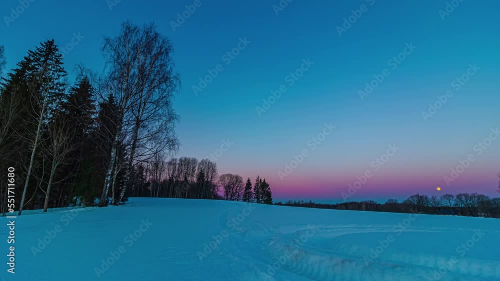 Poster blue hour timelapse over snow covered field with trees and setting moon in the sky in the distance. 