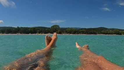 First-person water surface perspective of woman and female child legs and feet relaxing while floating on sea water of Santa Giulia beach in Corsica island, France. Low angle slow motion at 50fps