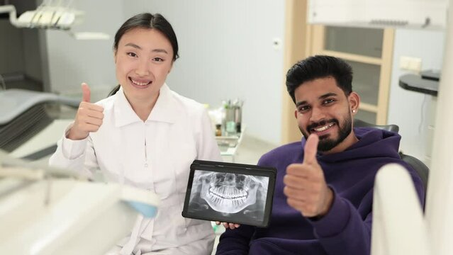 Smiling happy bearded man visiting dentist, sitting in dental chair at modern light hospital clinic. Young asian woman dentist holding tablet pc with x ray scan.