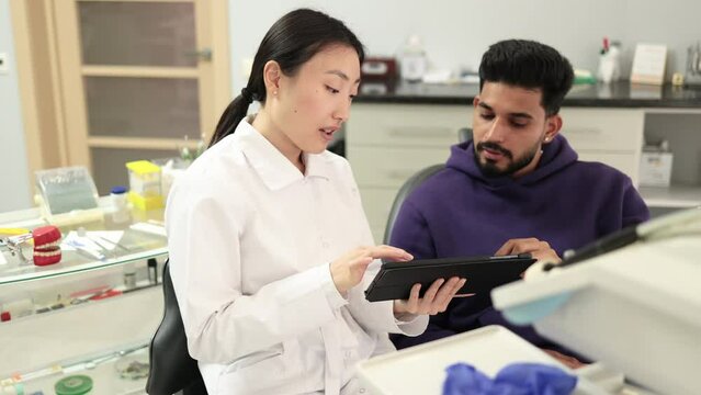 Medicine, dentistry and oral care concept. Female confident asian dentist showing tablet pc computer to worried male patient, bearded young man at modern dental clinic.