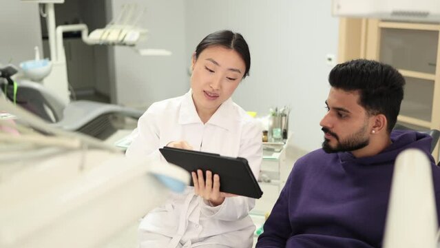 Medicine, dentistry and oral care concept. Female confident asian dentist showing tablet pc computer to worried male patient, bearded young man at modern dental clinic.