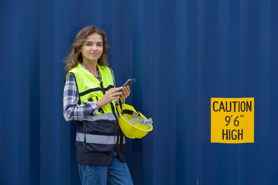 Young caucasian woman in hardhat and safety vest typing text massage on mobile phone. A large cargo container is in the background - Powered by Adobe