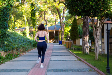 A female athlete runs along the sidewalk in the hotel along green palm trees, firs and pines.