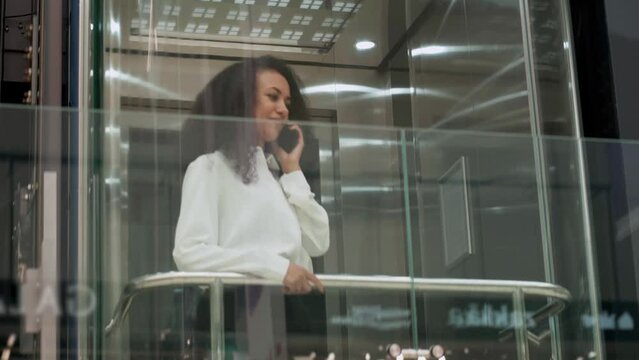 young woman talking on a smartphone standing in an elevator with a glass wall.