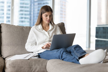 Focused young adult student girl using laptop at home, resting on soft couch at heap of papers, typing. Blogger writing essay, article, chatting, using computer for online communication