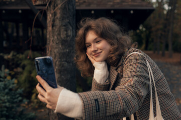 Young smiling girl walking in autumn park taking selfie pictures using smartphone.