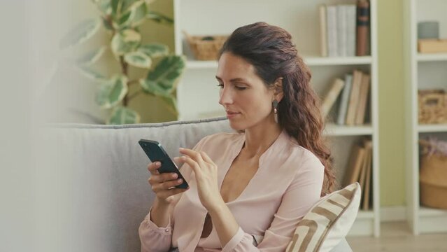 Young beautiful Caucasian woman with long brown hair scrolling on smartphone while sitting on soft couch in contemporary living room
