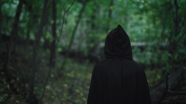 A dark figure in a black hooded robe walks through the mystical forest. Unrecognizable medieval witch.