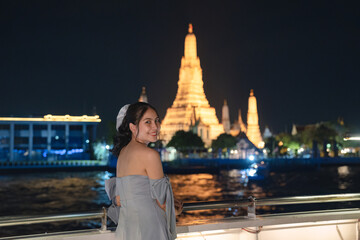 Beautiful young asian woman in dress standing and looking at camera on the deck with Wat Arun glowing in the night. Dinner cruise sailing on the Chao Phraya river - obrazy, fototapety, plakaty