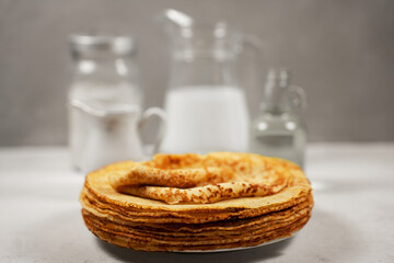 Round thin pancakes lie in a pile on a plate on a gray background with a jug of milk and containers with flour and sugar. Side view.