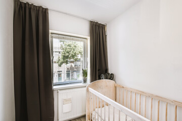 Interior of white room with baby crib and armchair near window with blue chest of drawers in...