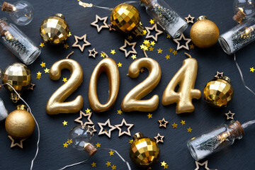 The golden figures 2024 made of candles on a black stone slate background are decorated with a festive decor of stars, sequins, fir branches, balls and garlands. Greeting card, happy New Year.