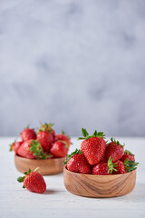 Ripe strawberries in two wooden bowls on a white wooden table on a gray background. Vertical, copy space, close-up.