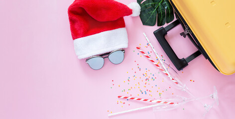 Christmas background.Flat lay.Yellow suitcase, Santa hat, paper straws, two glasses, sunglasses and confetti on pink background. Concept: travel in New Year holidays, vacation, tourist trips. Banner
