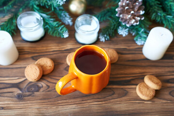 Orange coffee mug on a wooden background with white candles, oatmeal cookies and Christmas tree branches. The concept is a New Year's Eve photo wallpaper.
