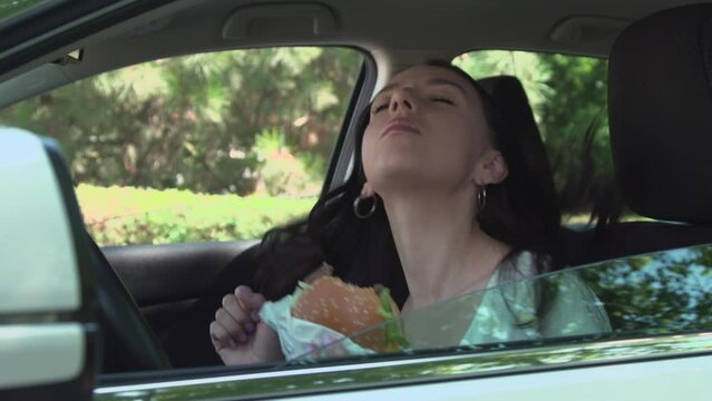 A young woman eating a hamburger and listening music in the car and saw friends and was shy. Fast food Humor concept..