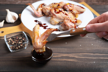The hand holds the fork with the chicken wing and dips it in the teriyaki sauce. Dish of Asian...