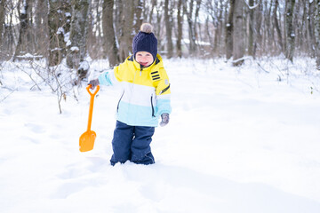 Cute smiling little boy with shovel playing with snow outdoors in winter forest