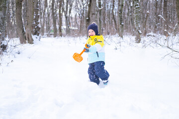 Cute smiling little boy with shovel playing with snow outdoors in winter forest