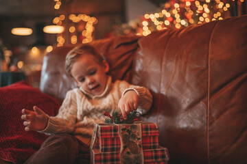 Candid authentic happy child in knitted beige sweater sitting with presents at lodge Xmas decorated