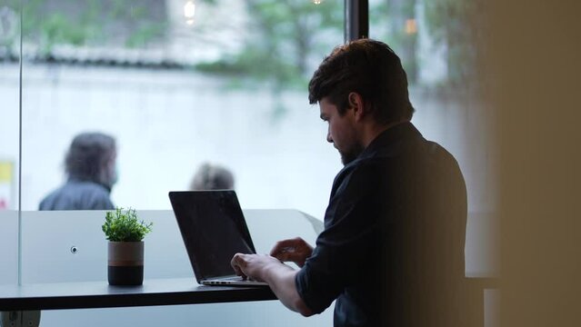 Candid young man works remotely with laptop at co working office typing on keyboard by window at urban street sidewalk