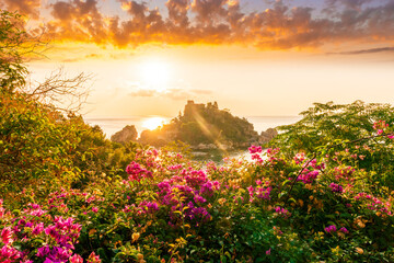 sunrise or sunset view to a  beautiful isle in sea from green and red flower bushes on foreground with clouds on the background of landscape