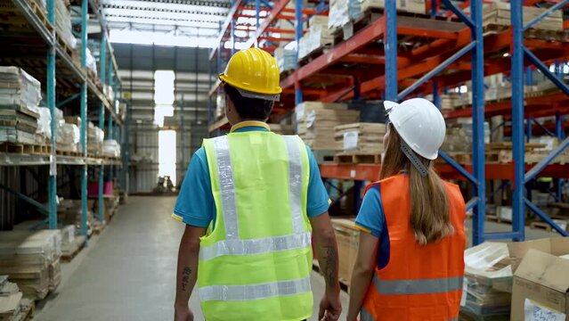 4K, Behind the back, two warehouse workers,  man and woman, are walking through shelves while inside warehouse. Two employees wear reflective suits and safety helmets to prevent accidents in warehouse