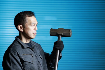 Craftsman in flayed work clothes holding a large metal hammer in front of the blue shutters of a town factory. Concept images of the essence of manufacturing and technical succession.