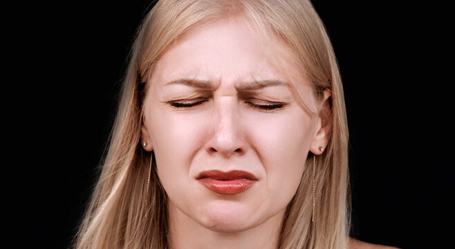 Headshot of young crying woman experiencing grief or loss. Grimace of heartache and closed eyes. Woman on black background. Girl is under severe stress and cannot restrain her emotions.
