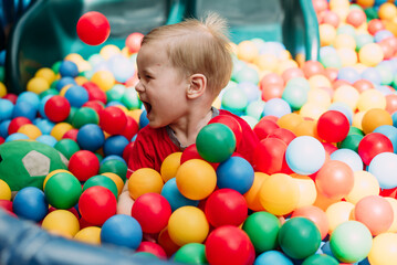 Fototapeta na wymiar Happy laughing boy 1-2 years old having fun in ball pit in kids amusement park and indoor play center. Activity toys for little kid