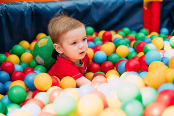 Fototapeta na wymiar Happy laughing boy 1-2 years old having fun in ball pit in kids amusement park and indoor play center. Activity toys for little kid