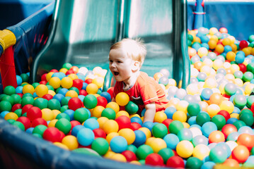 Obraz premium Happy laughing boy 1-2 years old having fun in ball pit in kids amusement park and indoor play center. Activity toys for little kid