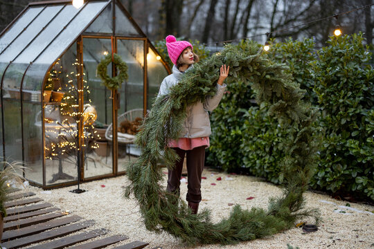 Young woman holds a big Christmas wreath made of pine twigs, preparing for a winter holidays at beautifully decorated and cozy backyard