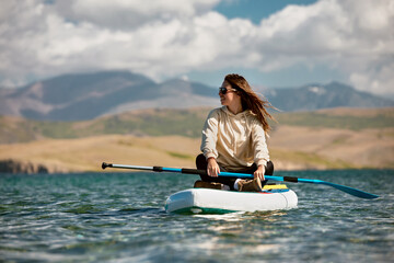 Happy young girl sits on sup standup paddle board at calm mountain lake