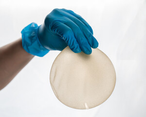 A female hand in gloves holds a teardrop-shaped breast implant on a white background. 