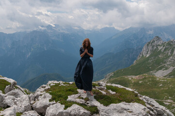 Naklejka na ściany i meble Beautiful young woman posing in a high mountain landscape. Rocky mountain. A young woman in a long dress stands on the edge of a cliff 