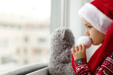 Sad alone child girl in Christmas red Santa hat looks at window and waiting holidays with teddy bear toy. Mental health