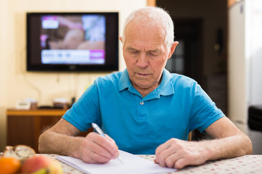 Serious Older Man Writing Letter At Table In Living Room