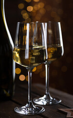Two glasses of white wine and a bottle on a wooden table and lights garlands behind
