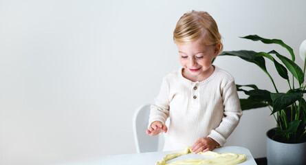Cute blonde serious girl,toddler playing with yellow sticky slime indoors at home on white...