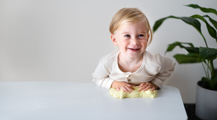 Cute blonde happy laughing girl,toddler playing with yellow sticky slime indoors at home on white...