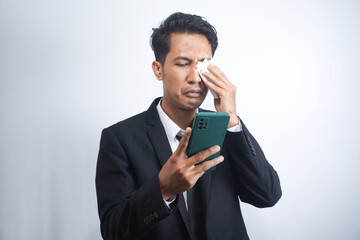 Asian guy wearing a suit looks sad reading online news from his cellphone. man shows disappointed gesture by wiping tears from his face