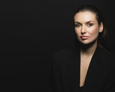 Fashion, business and make-up concept. Portrait of beautiful and sexy brunette woman with black suit in dark studio background. Model standing in grace pose and looking at camera with seductive look