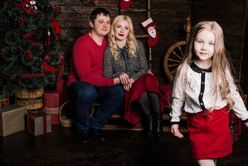 Obraz na płótnie Canvas Beautiful young family in red having fun together for Christmas holidays, sitting on a living room floor next to a nicely decorated Christmas tree