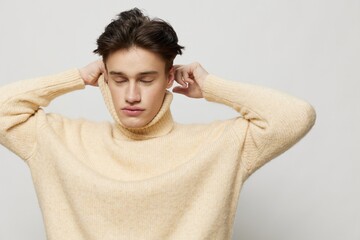 Horizontal studio shot. A beautiful young man with dark short hair combed back in a beige sweater with a high neck stands on a gray background